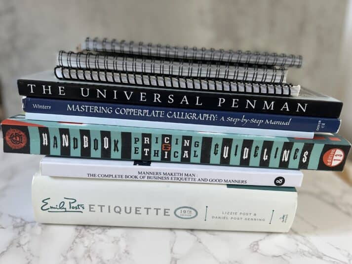 stack of calligraphy books with thick white on bottom, a blue striped spine and a black spine, topped with spiral bound books